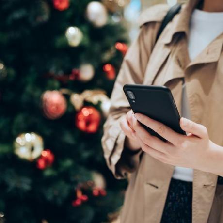 изрязан кадър и средна част на жена, използваща смартфон пред цветна коледна елха в празничния коледен сезон