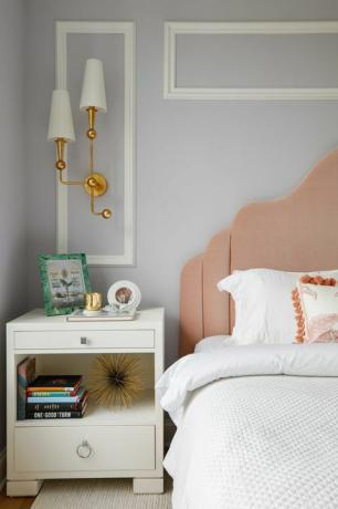 спалня, легло с бял лион, бяла масичка за легло, книги, розово табла, бели и златни аплици, бяла корона върху светло сиво боядисани стени