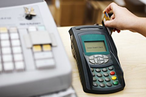 Ръката на жената прекарва карта през машина с кредитна карта, стояща до касов апарат. Фокусът е върху машината за кредитни карти.